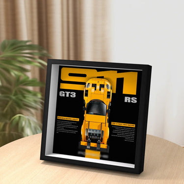 Framed art featuring a yellow Lego Porsche 911 GT3 RS model on a black and yellow background, displayed on a wooden table