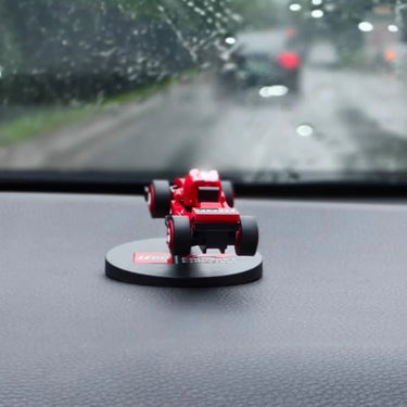 Red LEGO mini Formula 1 car model on a black round base on a car dashboard, rainy road visible through windshield